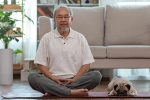 An elderly senior man relaxes with crossed legs and eyes closed doing yoga with a pug companion, dogs and seniors at assisted living community